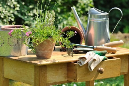 A close-up view of a wooden garden table situated outdoors, displaying various gardening tools and plants. The table holds potted herbs with green foliage, a metal watering can with a curved spout, and gardening gloves placed neatly on the tabletop. In the background, there are blurred green shrubs and trees, suggesting a well-maintained garden environment. The scene is captured in natural daylight, highlighting the textures of the wooden surface, the metallic sheen of the watering can, and the vibrant greenery, reflecting typical gardening activities in Kentish Town. Such an outdoor setting demonstrates an active garden management space, ideal for gardening tasks supported by local gardening services like those offered by Garden Maintenance Kentish Town.