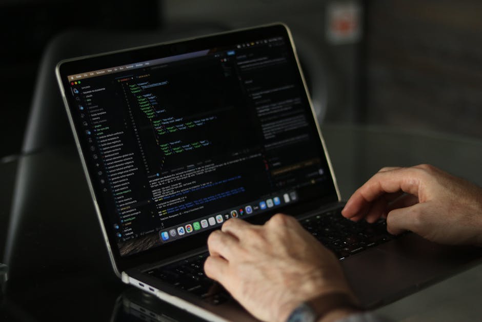 A person working on a laptop with a black keyboard and a bright, high-resolution screen displaying lines of colorful code in green, blue, and white against a dark background. The workspace appears to 