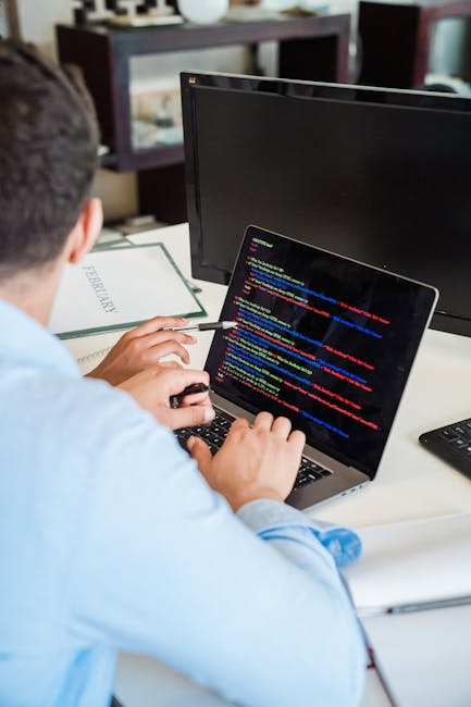 A person wearing a light blue shirt working at a desk with a laptop displaying colorful lines of code on its screen. The workspace includes an external monitor, a pen in the individual's hand, a noteb